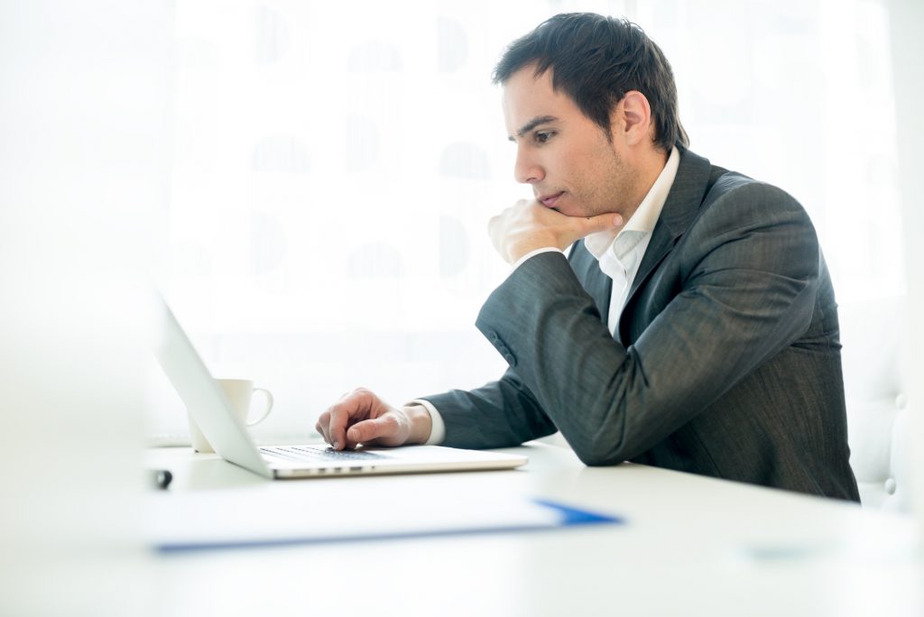 Serious young businessman concentrating on his work as he sits at his desk leaning on one arm navigating the internet on his laptop computer while reading the screen, profile view.